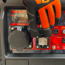 Mano enguantada manipulando una de las tomas de corriente del panel del generador, junto a puertos AC y DC, con pantalla digital visible al fondo.